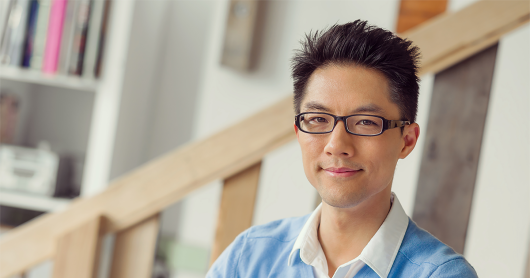 Young businessman in office wearing a play blue jumper over a white shirt. He has glasses on and he is smiling at the camera.