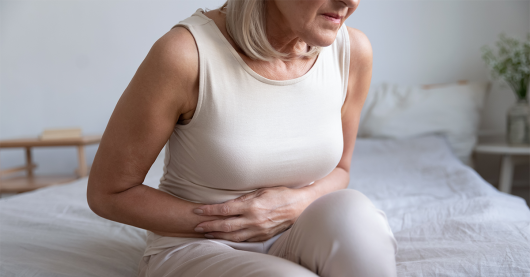 Senior woman sits on the end of a bed clutching her abdomen. She is wearing white trousers and a cream coloured sleeveless top.