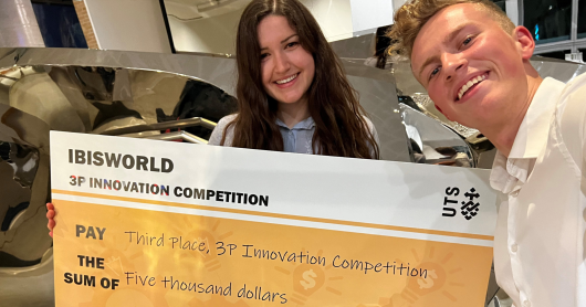 Thomas (right) takes a selfie with Agata, who is holding a large novelty check that reads "IBISWORLD 3P Innovation Competition". Agata is brunette with wavy hair and Thomas is blonde with a short cropped hairstyle. 