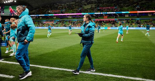 A woman dressed in a teal uniform holds a clipboard on the sidelines of a football pitch, players training in the background.