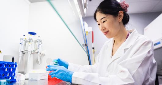 Dr Jiao Jiao Li is wearing a lab coat and sitting at a desk with a microscope and other lab equipment.