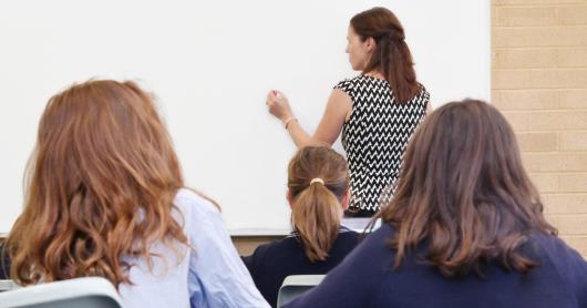 Three young students sitting in class look towards a teacher writing on a whiteboard.