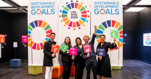 Five members of the UTS BUILD and Sustainability programs stand smiling in front of three vertical banners that read “SUSTAINABLE DEVELOPMENT GOALS”. The banner features multicoloured circular graphics. The members hold orange, green, pink and blue signs that correlate with the United Nations 17 Sustainable Development Goals.