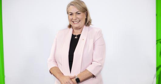 Dr Michele Bruniges AM stands against a white wall wearing a pink jacket and a black shirt, smiles at camera.