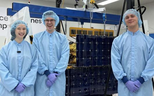 Engineering students Liv, John and Jacob wearing blue PPE gear standing next to satellite Optimus