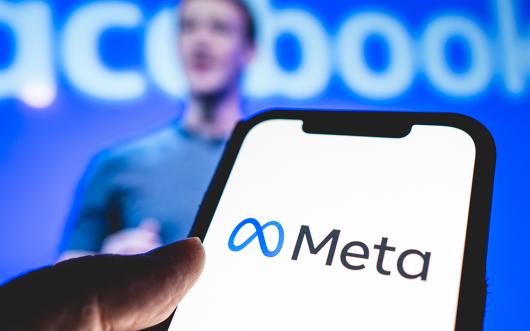 Strock picture of the Meta logo displayed on a mobile phone with an out of focus Mark Zukerberg in the background. 