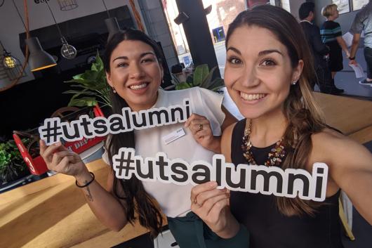 Two woman smiling and looking at the camera holding a #utsalumni sign