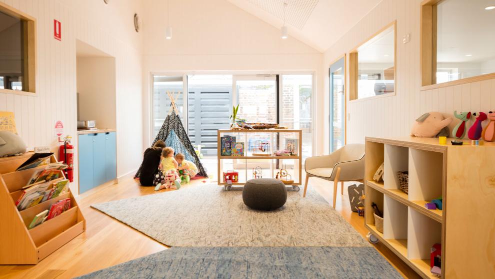 Two children ducking into a tent with an educator in an indoor setting at child care