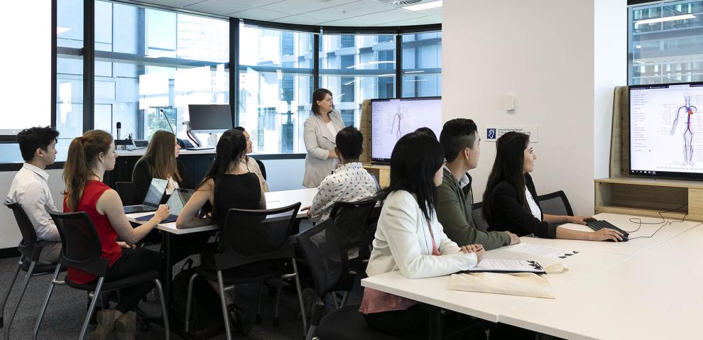 Groups of students work around tables with large screens at the ends while an instructor delivers information