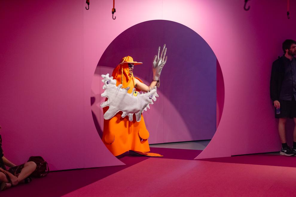 A person in an orange and silver costume emerges from a round doorway painted pink