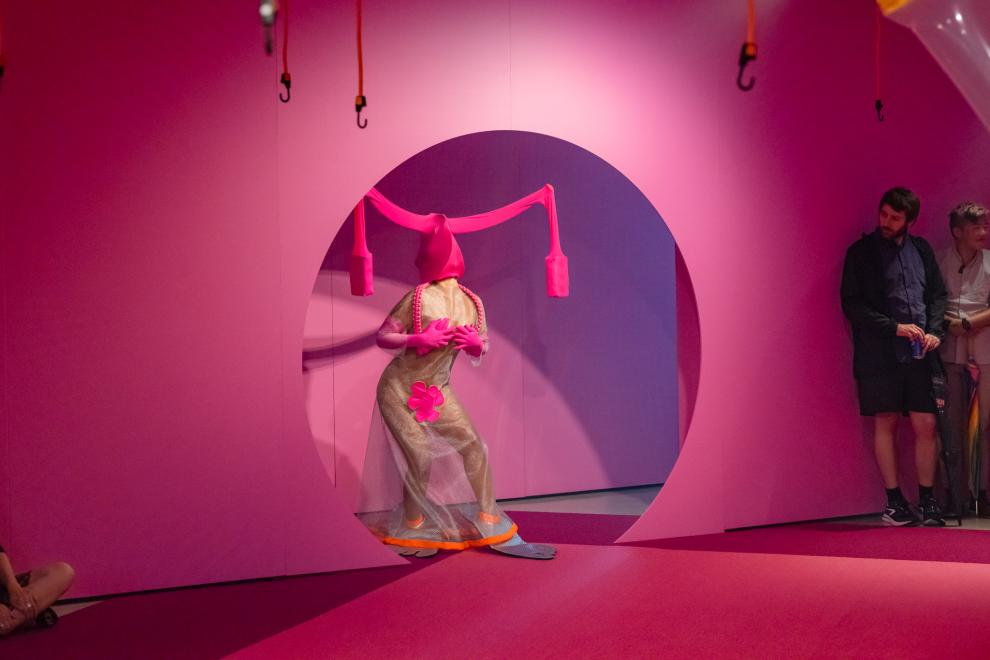 A person in a pink costume emerges from a round doorway painted pink