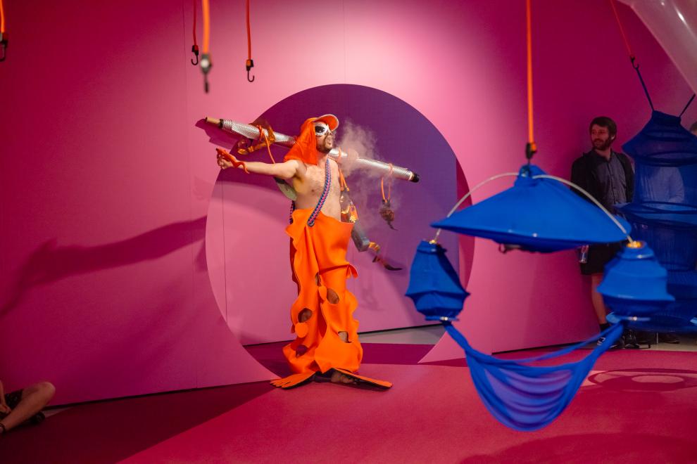 A person in an orange costume emerges from a round doorway blowing vape smoke
