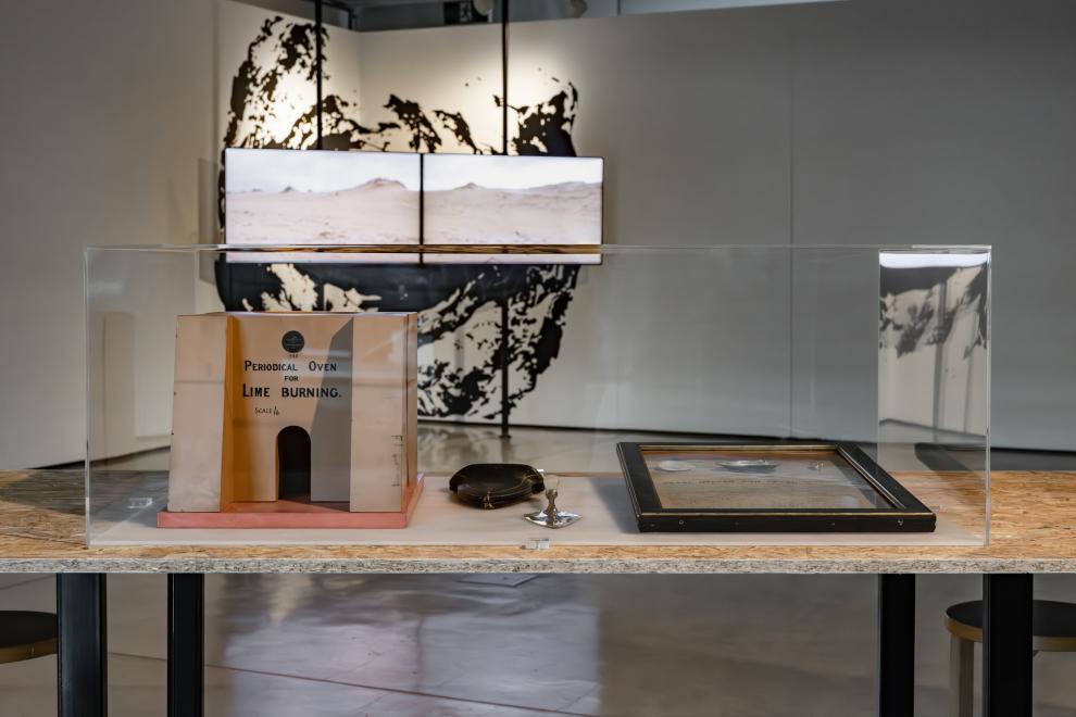 A clear acrylic box displaying objects is seen in front of two screens. 