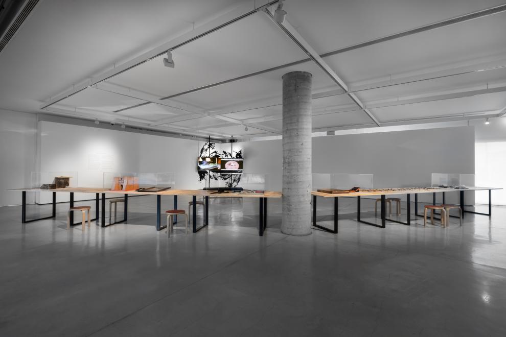 A gallery with a concrete colum in the centre. A table spans either side of the column. On the table are acrylic cases with objects inside. Two screens can be seen in the background. 