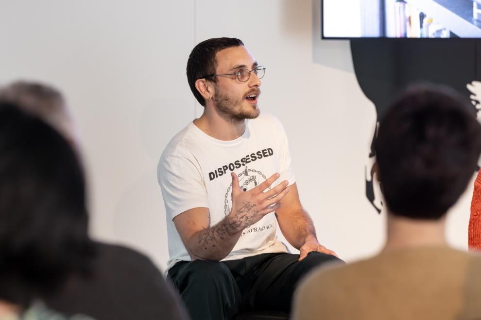 A person wearing glasses and a tsirt with the word "dispossessed" on it addresses a crowd 