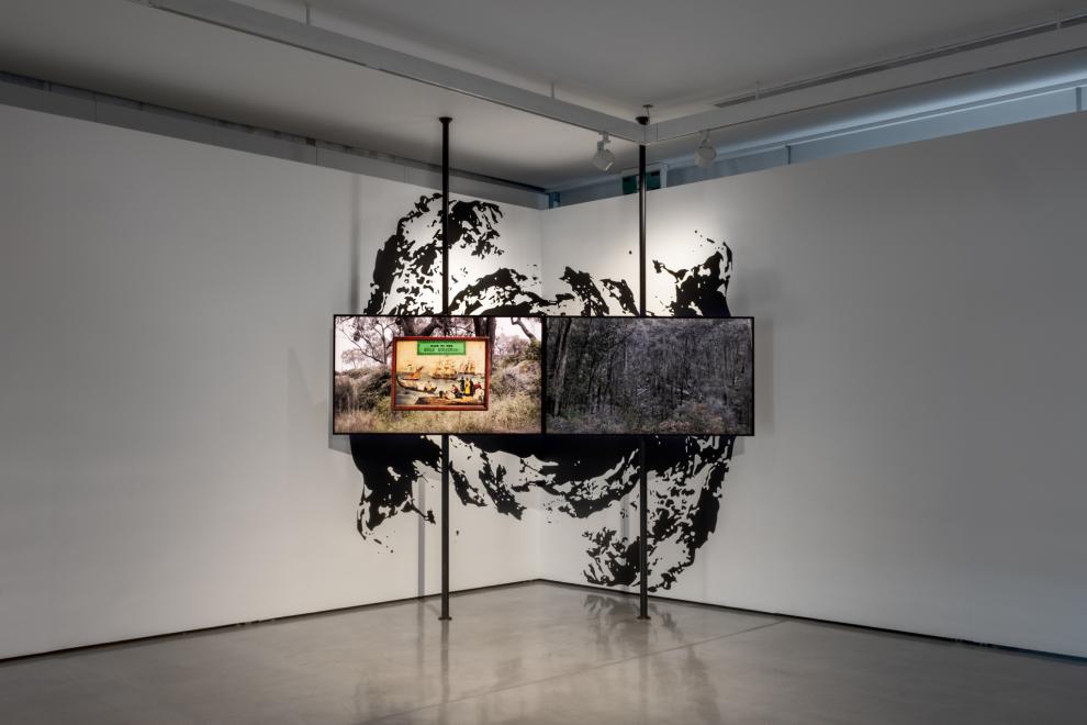 Two television screens mounted on vertical poles. The screen on the left shows a game box, the screen on the right shows a landscape. Behind the screen a black and white graphic of a mine shaft is on the wall. 