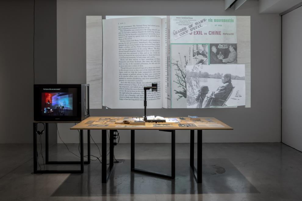 a sculptural installation by Prima Jalichandra-Sakuntabhai, which comprises a large timber table with carefully placed archival materials and a digital document camera which is projecting a collage of these materials onto the wall beside the table. There is a smaller table of the same height and materials which holds a heavy 1990’s-era TV monitor with headphones on a hook below the screen