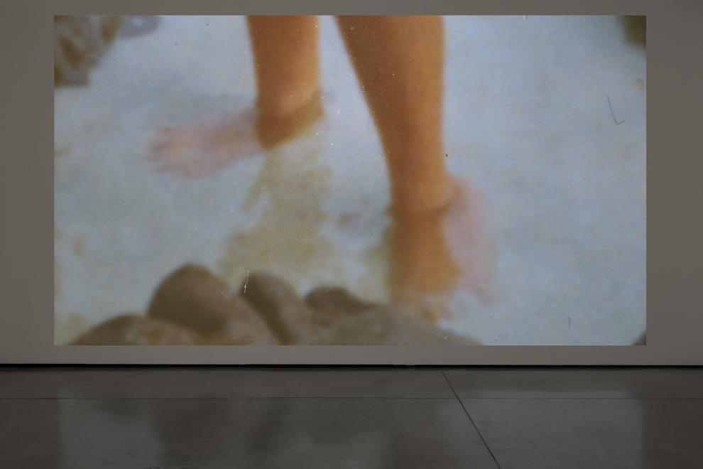 A photograph of a video installation which shows a person's feet in shallow water.