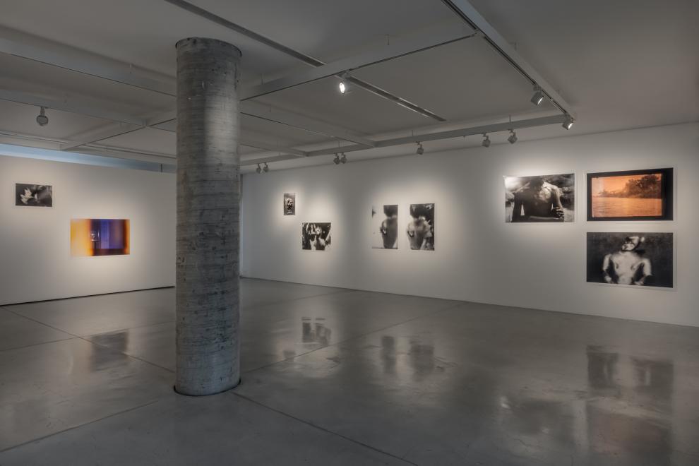 On the far wall of the main gallery space are 3 groupings of images. The right grouping are 3 large scale photographs of a pink-washed river scene, a mud-covered torso where the identifying head has been cropped out of the image, and a museum close-up of a burnished gold figurine from antiquity, that is blurred and falling into shadow. 