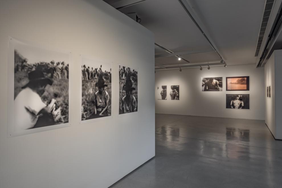 Photograph of a gallery installation showing a series of 3 slightly blurred black & white photographic portraits of men in cowboy hats, pictured in a field with a group of people gathered in the distance behind them. The images have visible crinkles and dust as part of the photograph.
