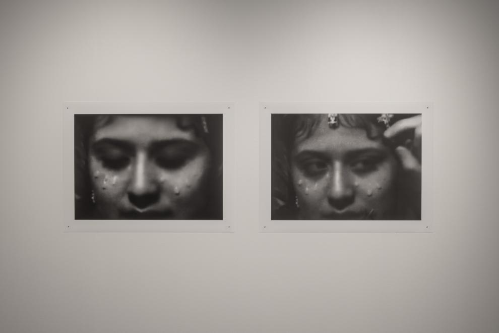Photograph of a gallery installation showing a pair of black & white photographs are pinned, showing slightly different close-ups of the artist’s decorated face. There are a series of teardrop adornments under the eyes and metallic charms hanging from the forehead in each image.