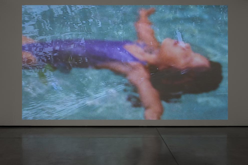 A photograph of a video installation which shows a small child floating on their back in a swimming pool.