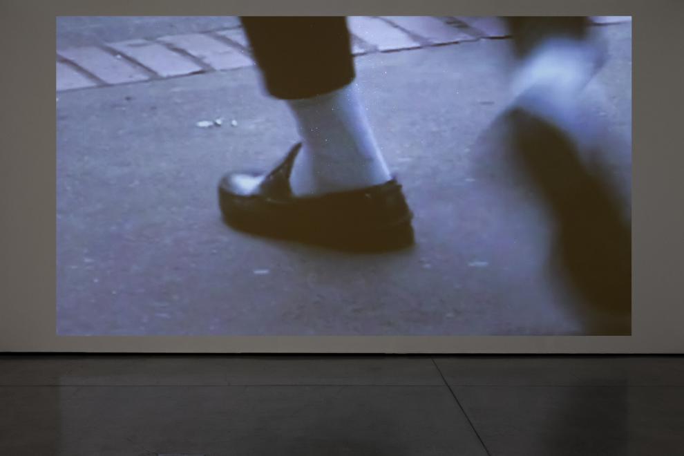 A photograph of a video installation which shows a person's feet in black loafers and white socks.
