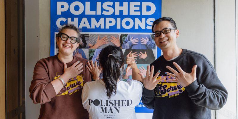 Two people are holding their hands up to show a blue nail, between them, one person has their back to the camera to show their shirt that reads: Polished Man. 