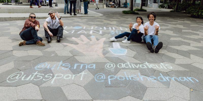 Four people on the ground, on the ground is chalk written messages about the Polished Man campaign. 