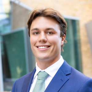 A portrait of Darcy Davies, he's smiling at the camera dressed in a blue suit and standing in front of the the UTS Business School.