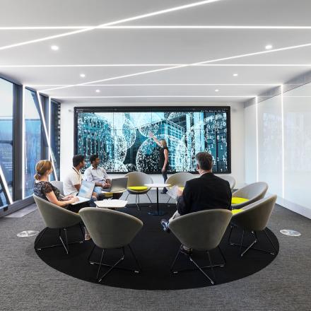 Photo of the UTS DataLounge. Four people are sitting in a circle of chairs looking towards the LCD screen where another person is presenting.