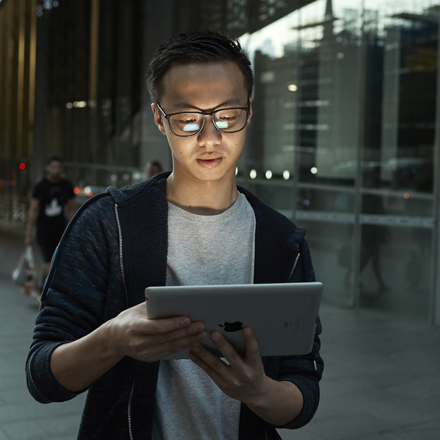 A young man stands on a street corner, he holds a laptop in his hands and is looking at the screen.