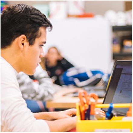 Secondary male student working on a laptop in a classroom with  female student looking on in the background