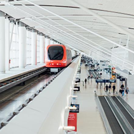 Red shiny train pulling up at a white indoor train station with people walking off travelators on the lower level