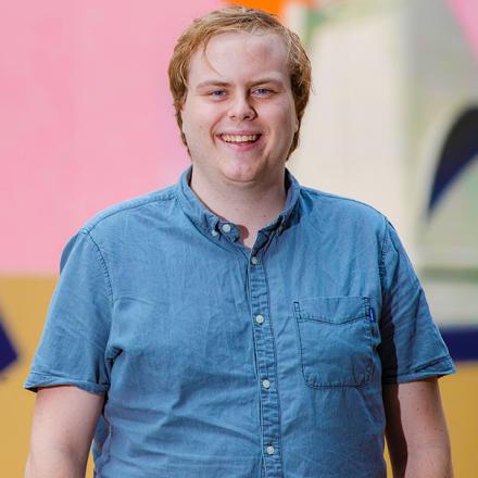 Young man smiles in front of pink mural