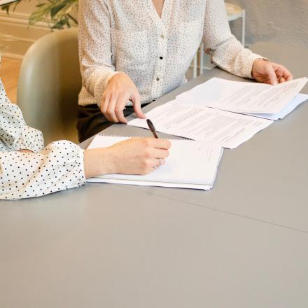 A photograph of two women at a table, depicted with their faces out of frame. One is writing and the other is shuffling papers.