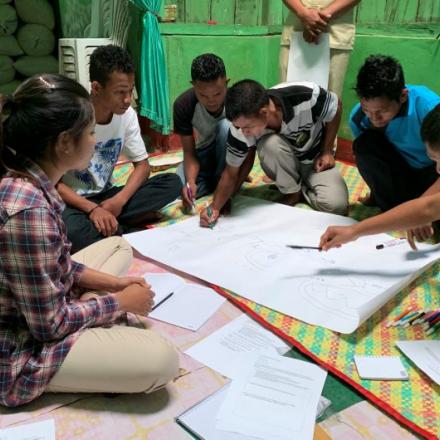 A group of men and women sit on the floor around a large piece of paper. Some are writing on it.