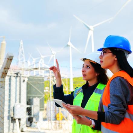 Two women in hi-vis gear stand in front of wind turbines. 