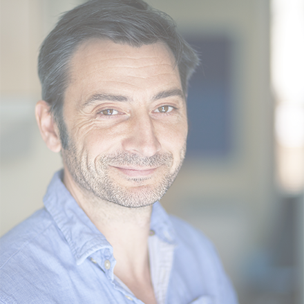 Dark haired man wearing blue shirt with open collar smiling at the camera