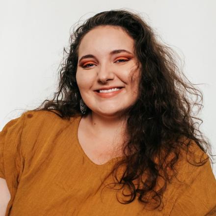 woman with long dark curly hair, burnt orange top, orange eye shadow, smiling at camera