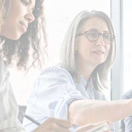 mature woman with shoulder-length grey hair and glasses is wearing a blue shirt. She is siitting beside a young woman with long brown curly hair in a grey and whitee striped shirt. The older woman is pointing at a computer monitor and the young woman is taking notes with a pen.