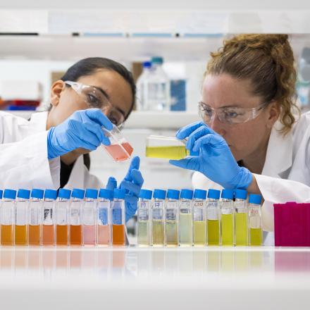 Two female scientists in white coats, blue gloved and goggles inspect a test tube containing orange liquid.