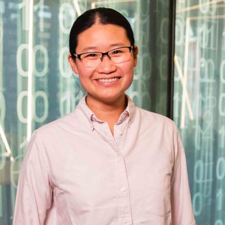 Jaime standing in front of a screen. She is wearing glasses and a bone coloured business shirt. She is smiling and looking at the camera.