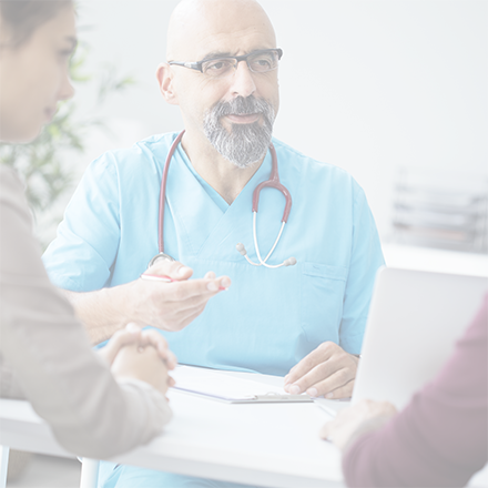 Doctor in blue scrubs with stethoscope around neck talks to patients at a desk