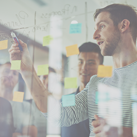 A group of people collaborating behind a glass panel covered in writing and sticky notes.lding a marker