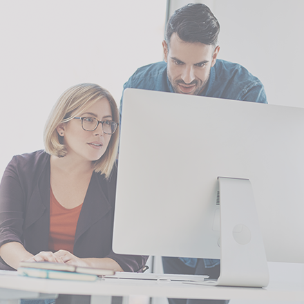 A woman seated at a desk and a man standing next to her. Both are looking at a computer in front of them