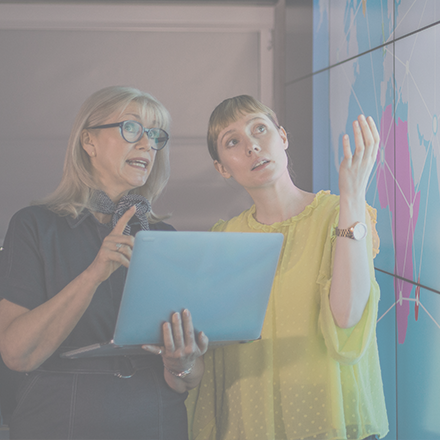 Two women in conversation, both are looking at a screen displaying a world map. One woman wears glasses and holds a laptop. The other wears a watch and a yellow blouse. 