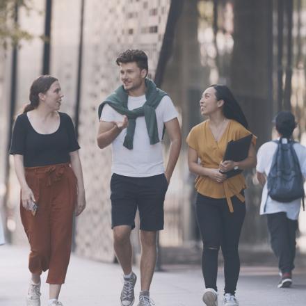 Three young adults walk and chat on the UTS campus. They appear relaxed and engaged, with modern glass buildings in the background and trees on the side.