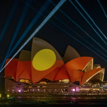 The Sydney Opera House is illuminated at night with vibrant red and yellow Indigenous flag art projections. Blue laser beams radiate into the night sky.