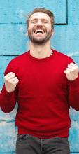 Man wearing red long-sleeved shirt standing beside wall with head up, eyes close and big smile with fists clenched in victory